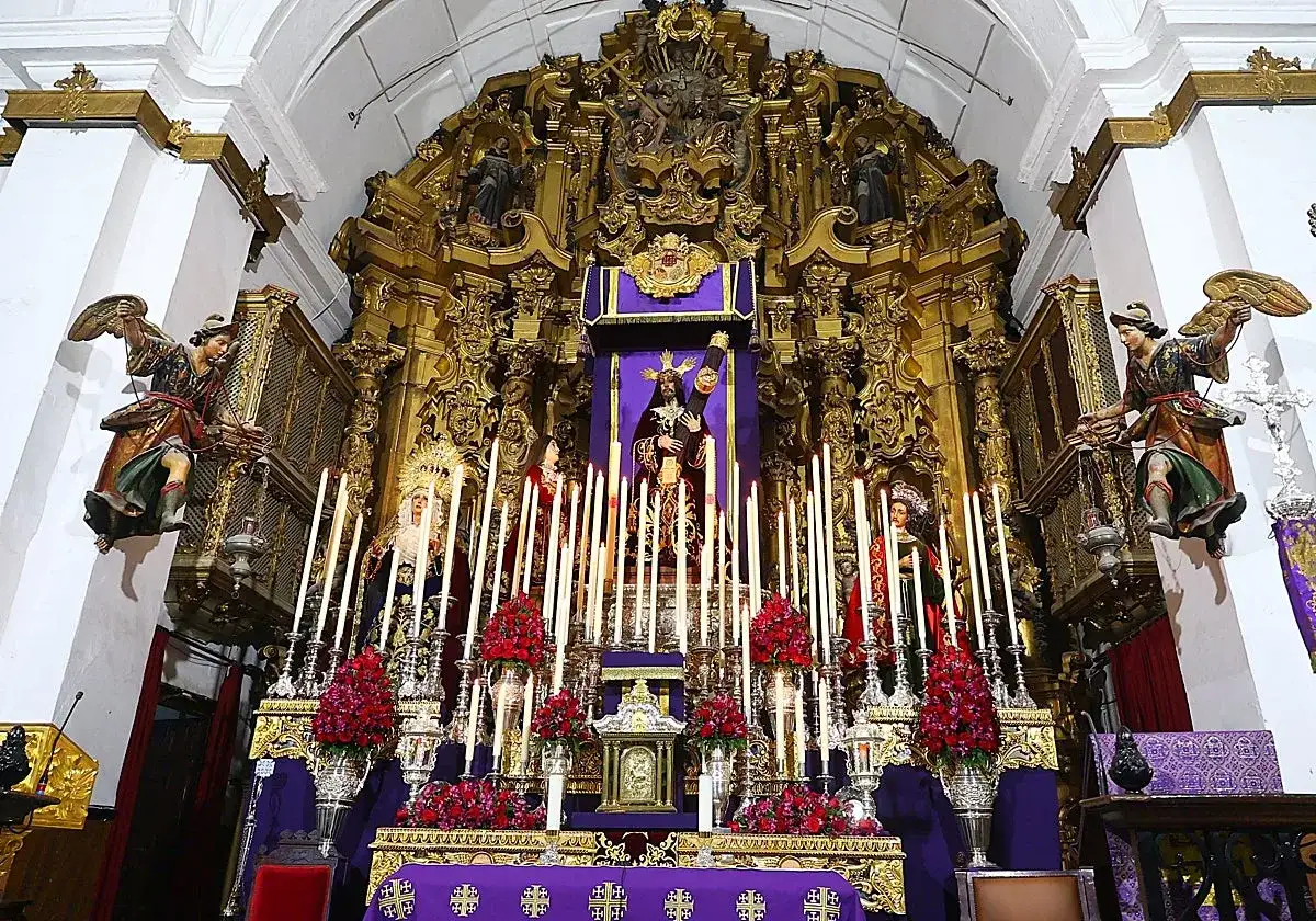 Un altar barroco con imágenes religiosas, velas y flores rojas. Destaca una figura central de Cristo, que parece ser un quinario, rodeado de figuras de la Virgen y otros santos.