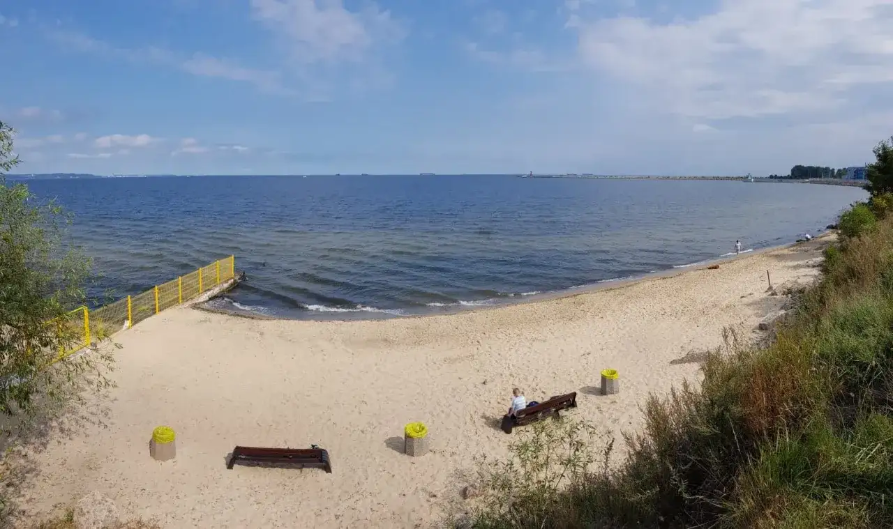 Plaża dla psów z łagodnym zejściem do morza. Na piasku ławki i kosze. W oddali widać falochron.