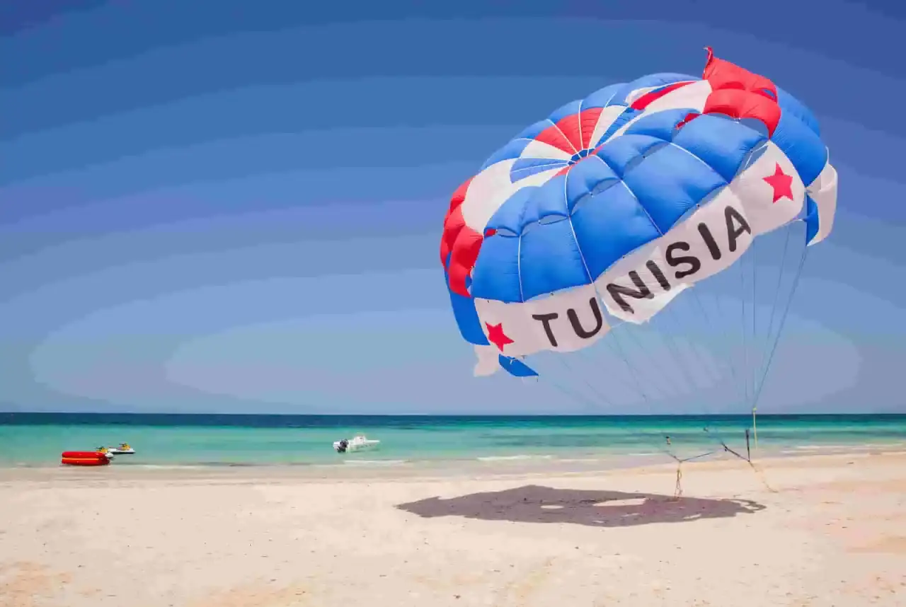 Kolorowy spadochron z napisem "TUNISIA" unosi się nad plażą, zapowiadając wspaniałe dżerba loty. W tle turkusowe morze i piaszczysta plaża.