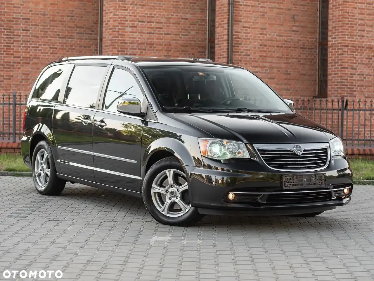 Czarny Lancia Voyager stoi na brukowanym podjeździe przed ceglanym budynkiem.