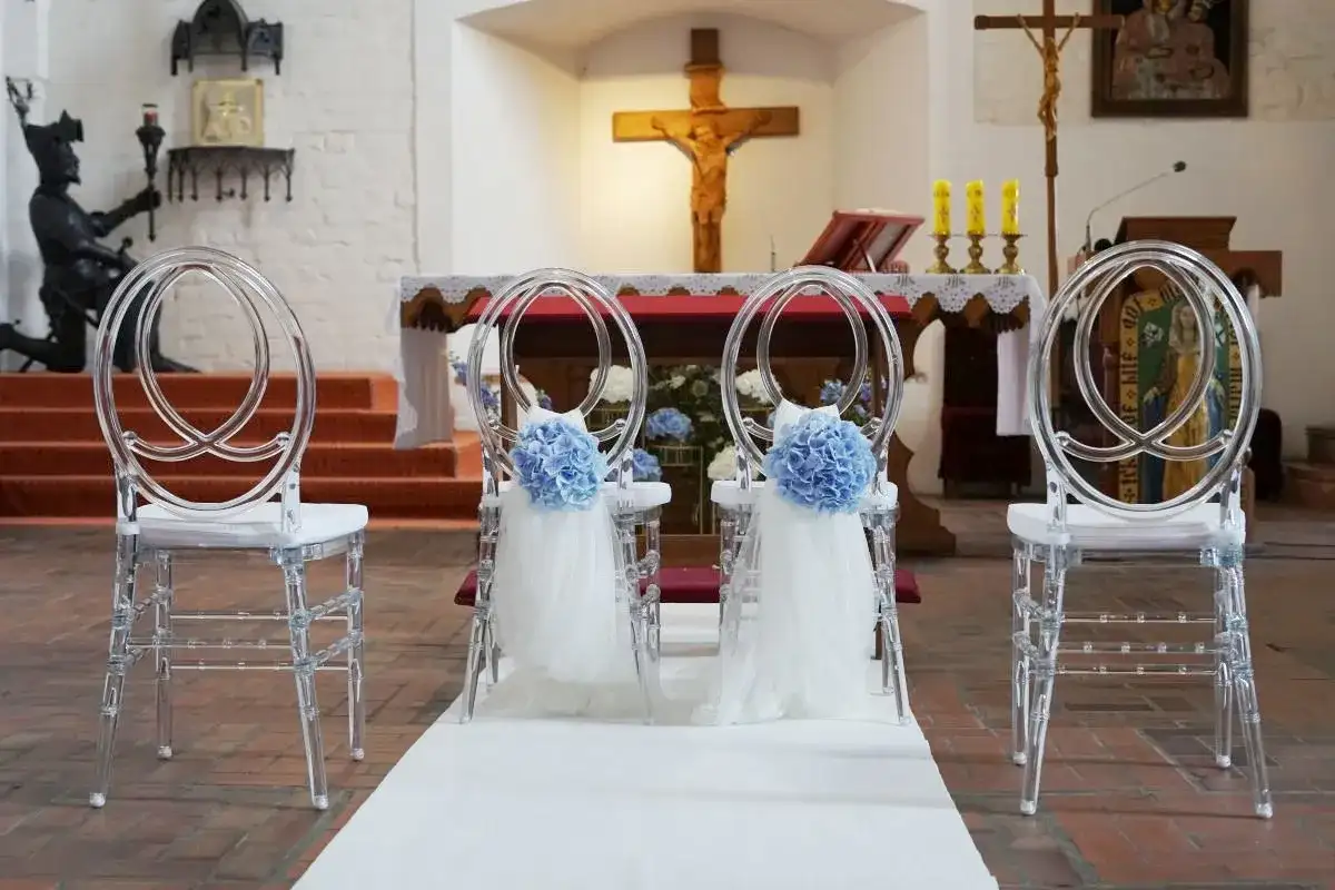 Elegancka dekoracja kościoła na ślub samemu: przezroczyste krzesła z błękitnymi kwiatami i tiulem, biały dywan prowadzący do ołtarza.