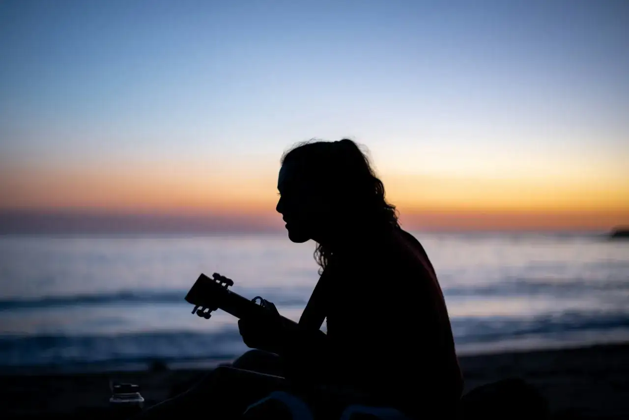 Sylwetka grającej na ukulele osoby na tle zachodzącego słońca nad morzem. Gdzie ta keja chwyty?