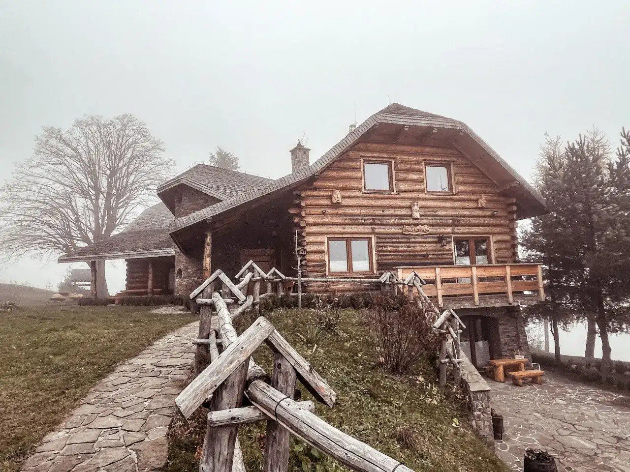 Drewniany dom z kamienną podstawą, kamienista ścieżka i płot. Mglisty dzień dodaje uroku temu miejscu, które oferuje liczne laskowa atrakcje.