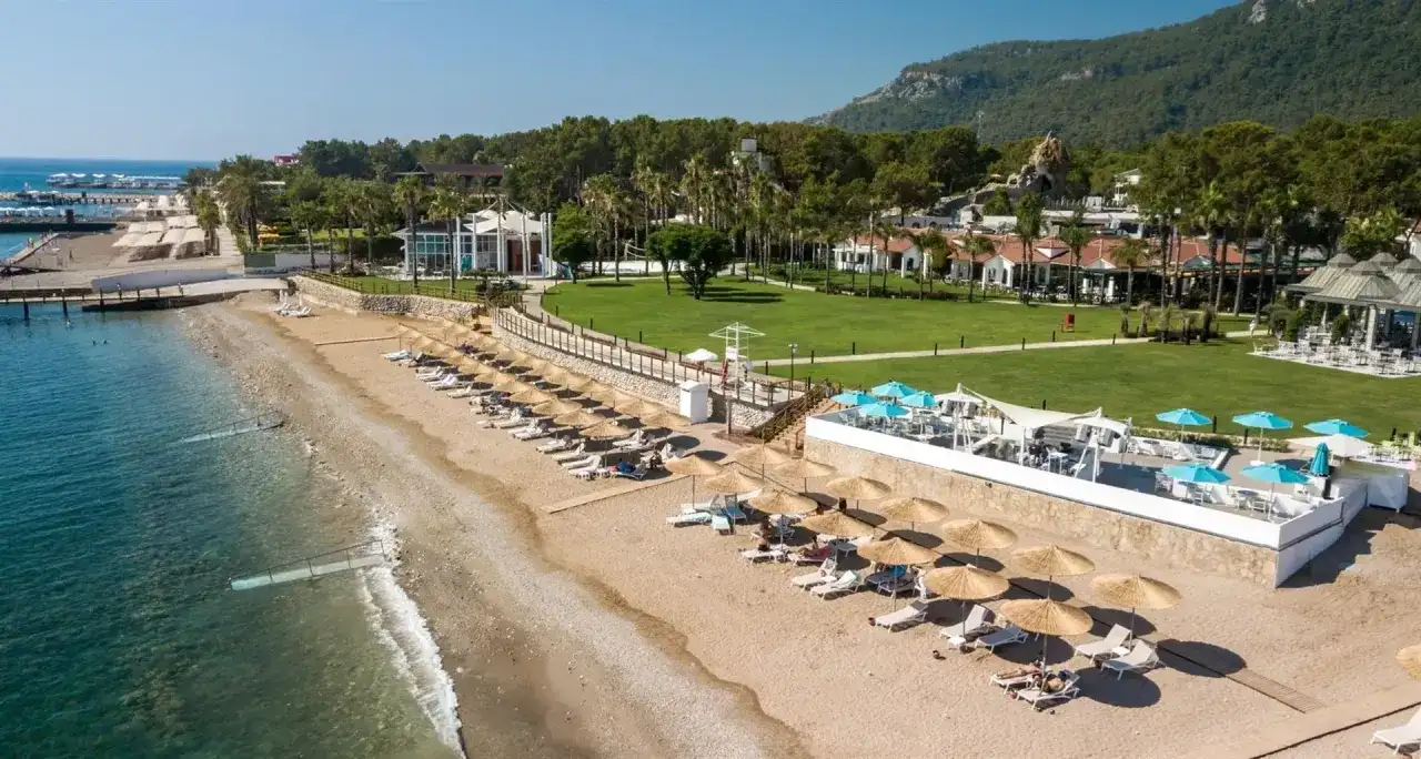 Słoneczna plaża Baia Salima Kemer z leżakami i parasolami, zielony trawnik hotelowy i góry w tle.