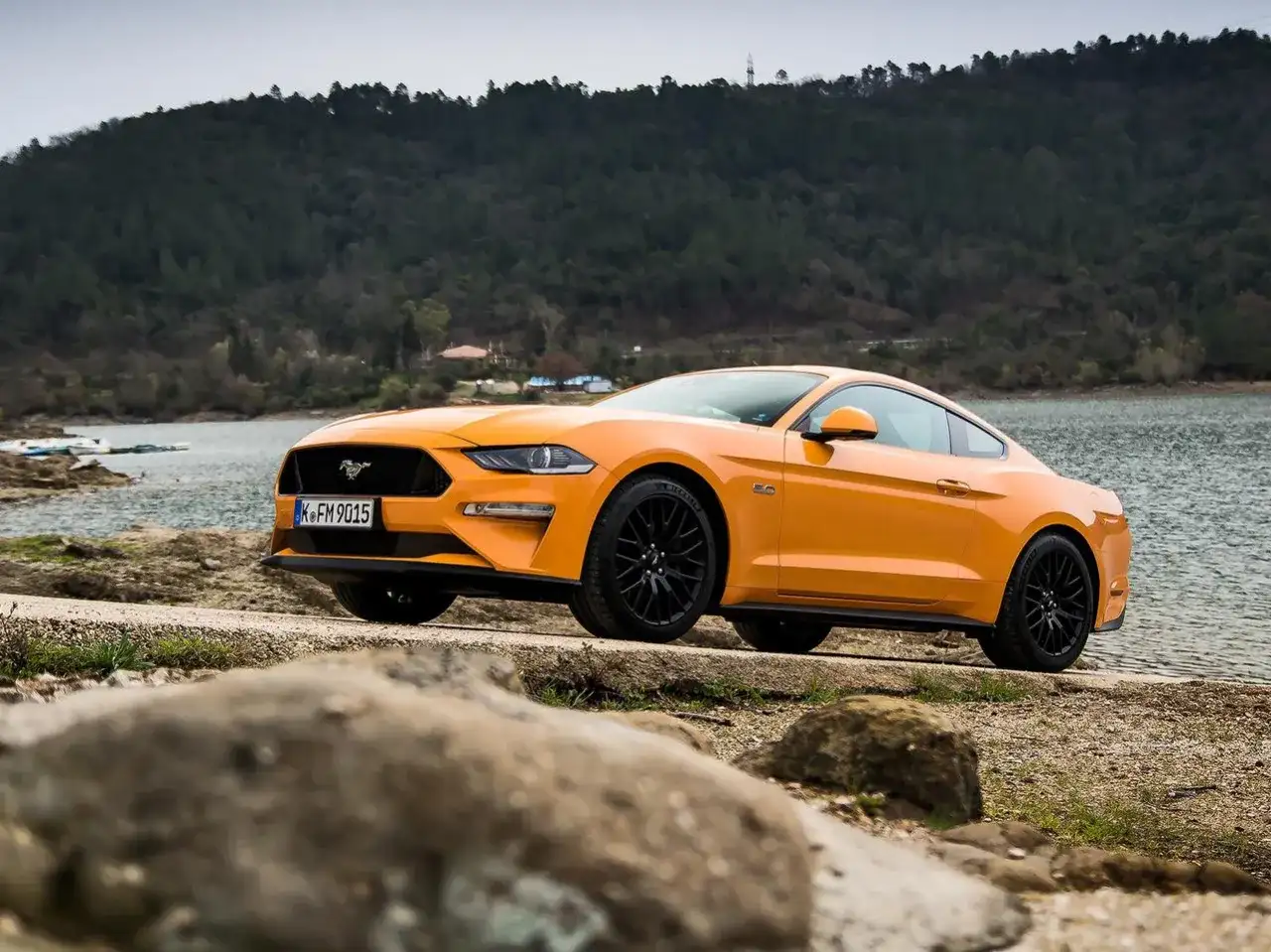 Pomarańczowy Mustang 2.3 Ecoboost z czarnymi felgami stoi nad jeziorem, gotowy na przygodę.