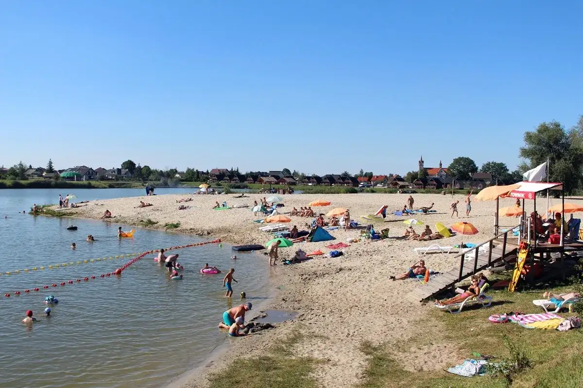 Słoneczny dzień na piaszczystej plaży bobrowego rozlewiska w Zabierzowie Bocheńskim. Ludzie opalają się, kąpią i bawią w wodzie.