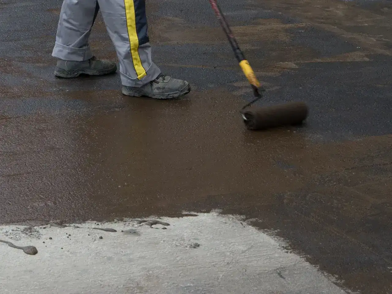 Pracownik w szarym ubraniu z żółtym pasem nakłada wałkiem ciemną masę na betonową powierzchnię. To zbrojenie rozproszone, które wzmacnia konstrukcję.