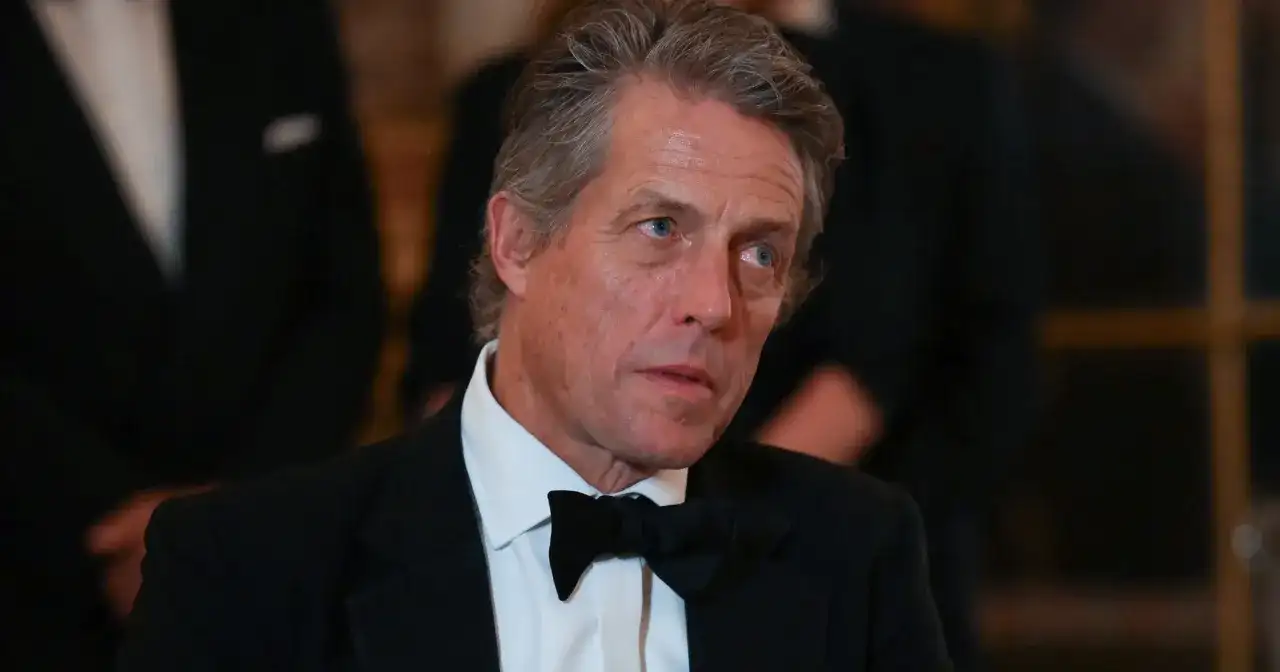 Hugh Grant w czarnym smokingu i muszce, z siwiejącymi włosami, wygląda na zamyślonego.