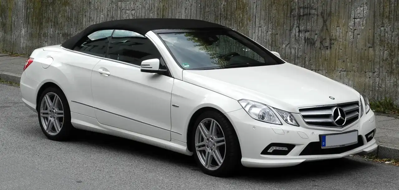 Biały kabriolet Mercedes W207 z czarnym dachem stoi na ulicy obok betonowej ściany.