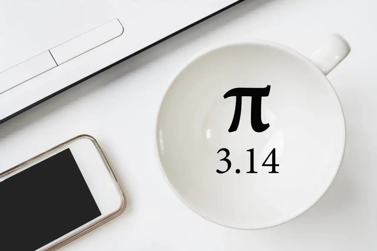 Biały kubek z symbolem π i liczbą 3.14, obok smartfon i fragment laptopa.