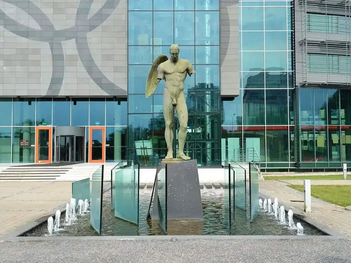Brązowe rzeźby przedstawiające postacie ludzkie, w tym anioła, stoją przed nowoczesnym budynkiem z przeszkloną fasadą.