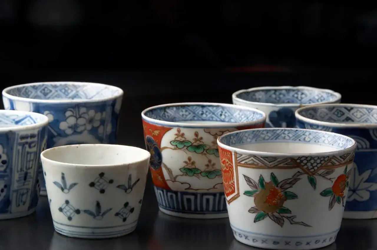 Vielfalt der japanischen Porzellankunst: Mehrere kleine Becher mit traditionellen Mustern, darunter Blumen und geometrische Formen, auf dunklem Hintergrund.