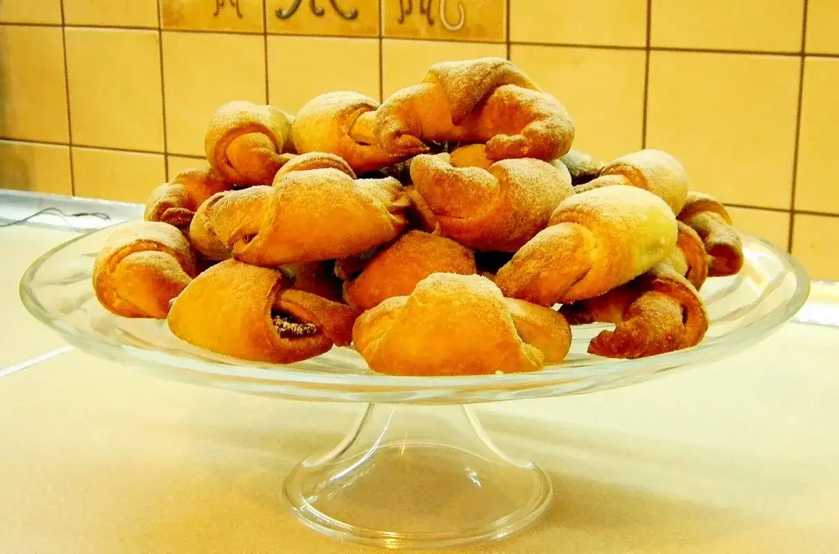 Rogaliki drożdżowe z przepisu babci, posypane cukrem pudrem, piętrzą się na szklanej paterze.