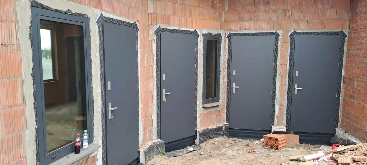 Nowe, szare drzwi zewnętrzne i okno w budowie. Widać ceglane ściany i materiały budowlane.