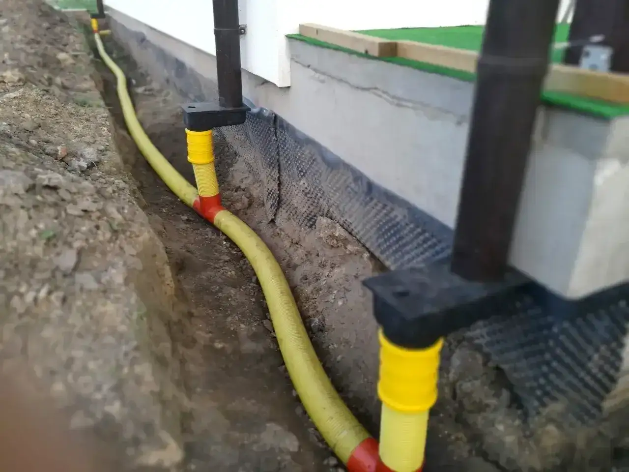 System odprowadzenia wody z rynny, z żółtą rurą biegnącą w wykopie przy fundamencie.