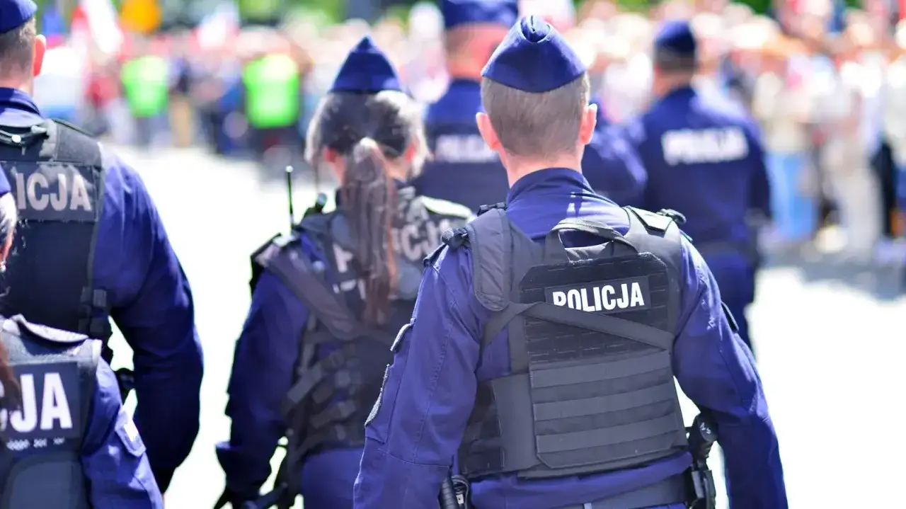 Policjanci w mundurach, tworzący zgraną kartę rodziny mundurowej, patrolują ulicę. Na kamizelkach taktycznych widnieje napis "POLICJA".