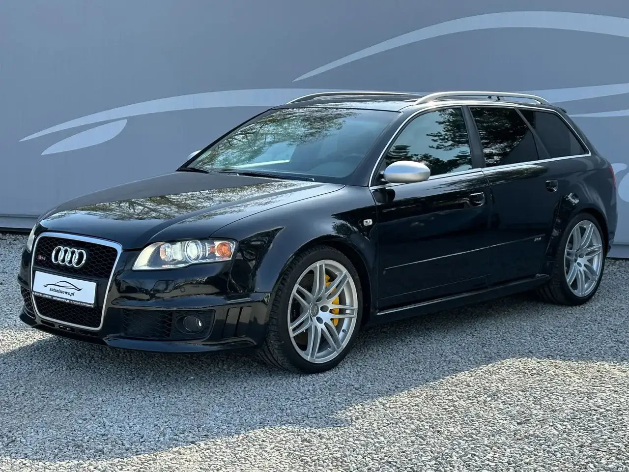 Czarny Audi RS4 B7 Avant z żółtymi zaciskami hamulcowymi, zaparkowany na żwirze przed szarą ścianą z grafiką.