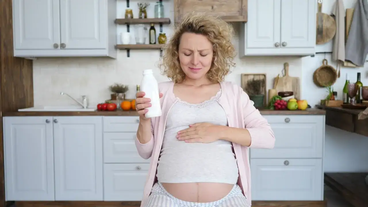 Kobieta w ciąży trzyma butelkę mleka, czując ból serca. Jej twarz wyraża dyskomfort, a dłoń spoczywa na brzuchu.