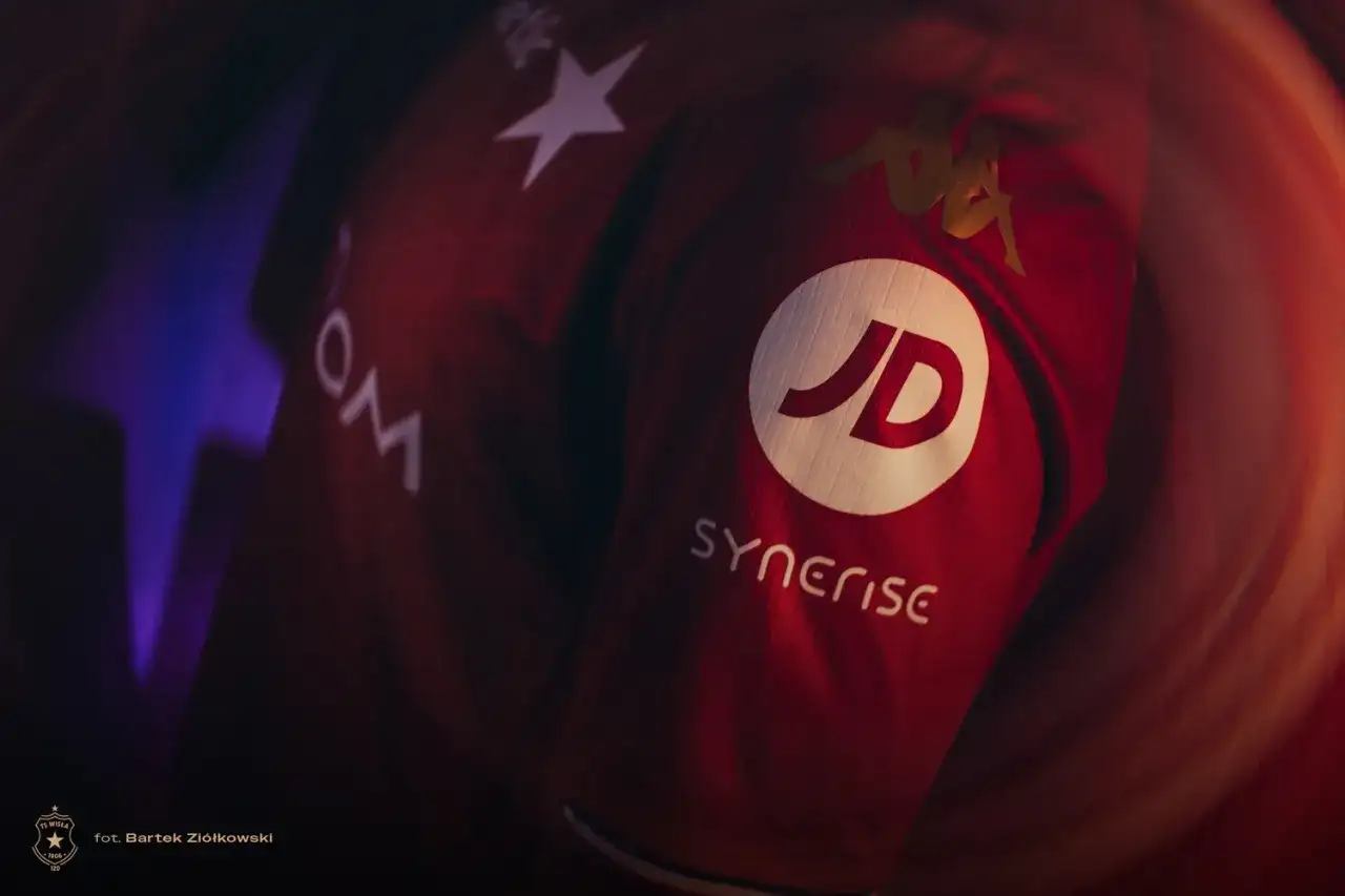 Czerwona koszulka Wisły Kraków z logo JD i Synerise. Widoczne też logo Kappa i gwiazda.