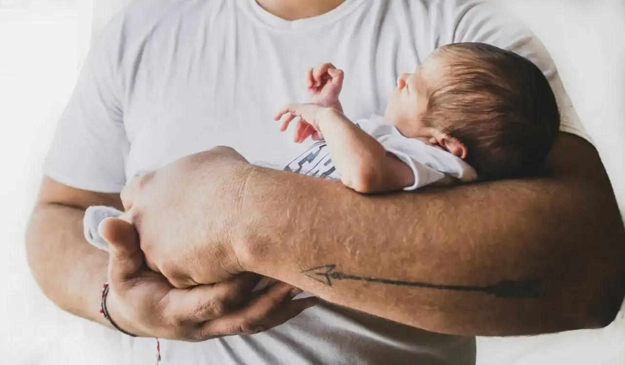 Ojciec trzyma dziecko, które wydaje się być w niewygodnej pozycji. Złe noszenie niemowlaka może być niebezpieczne.