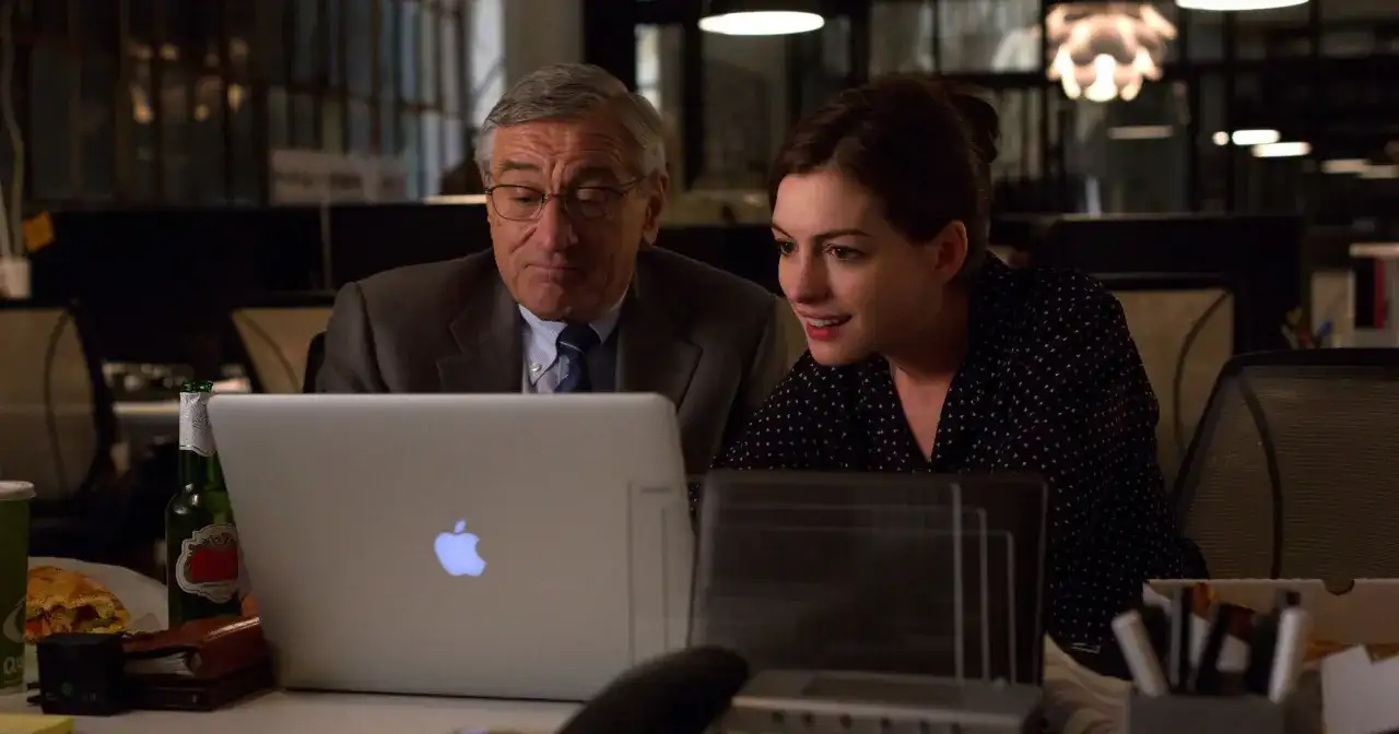 Robert De Niro i Anne Hathaway pracują przy laptopie. Młoda praktykantka z entuzjazmem analizuje dane, podczas gdy doświadczony szef czuwa nad jej pracą.