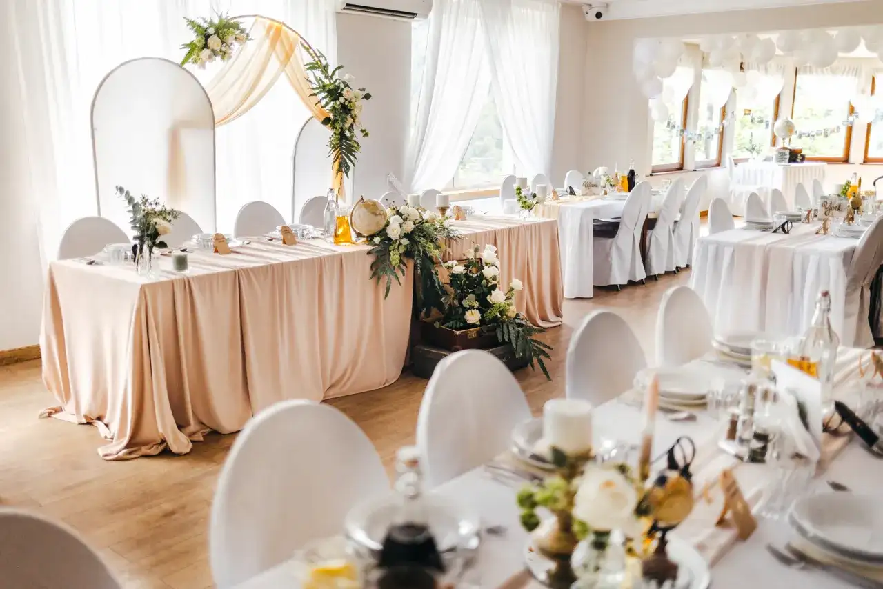 Elegancka sala weselna z białymi obrusami i kwiatami. Na stole pod dębem przywory stoi globus i walizka.