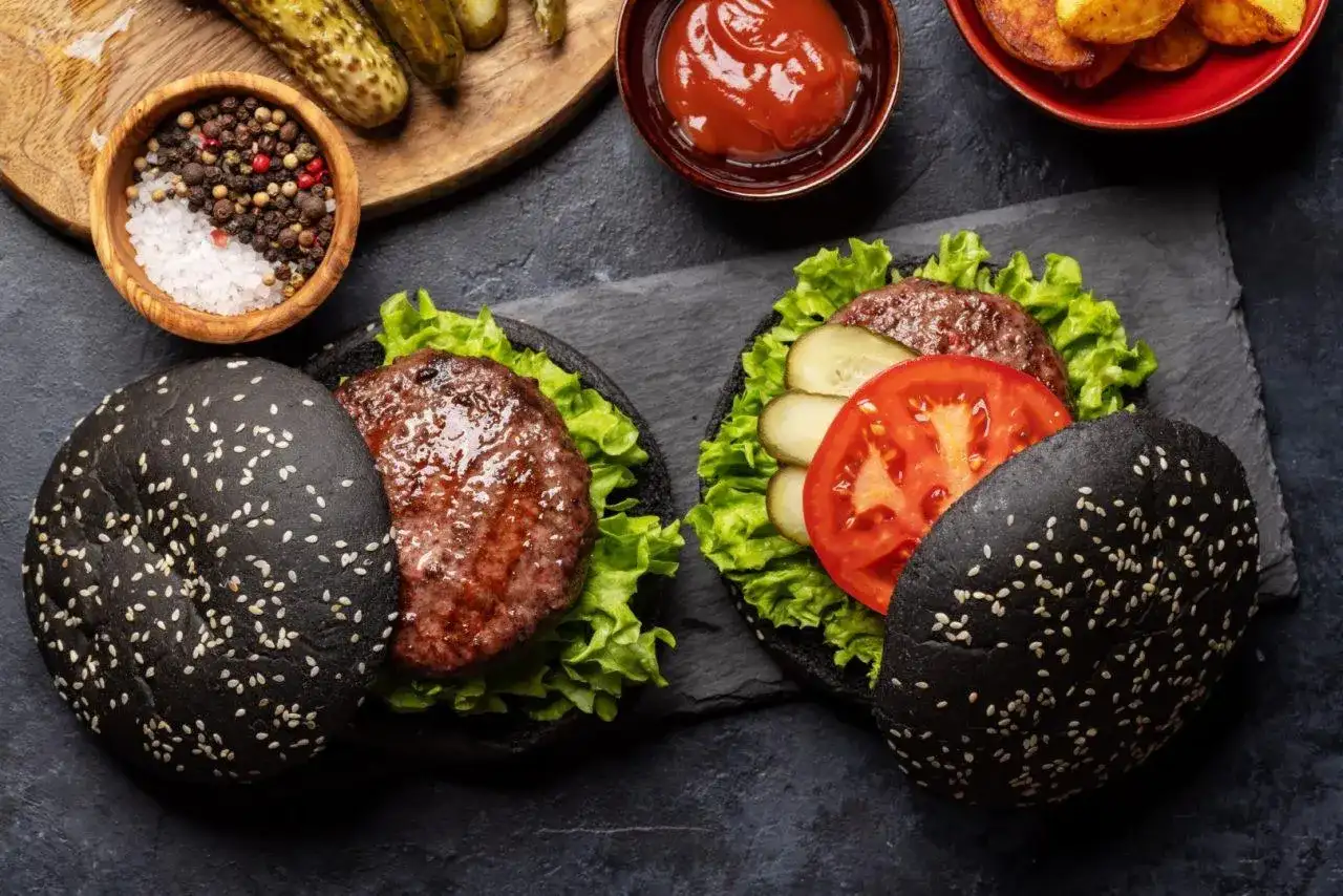 Dwa czarne burgery z sezamem, sałatą, pomidorem i ogórkiem kiszonym.