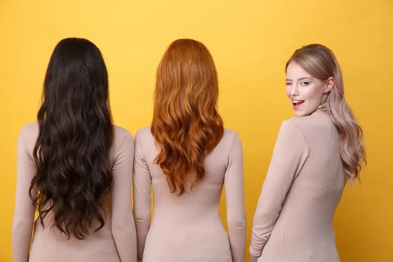 Trzy kobiety prezentują modne kolory włosów: kruczoczarne, rudoczerwone i pastelowy blond. Jedna z nich mruga do widza.