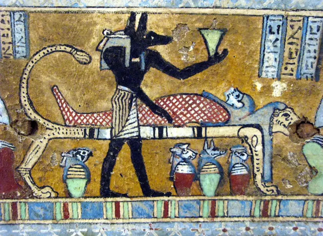 Anubis, bóg z głową szakala, stoi przy katafalku z mumią. W ręku trzyma naczynie, a obok stoją urny z głowami zwierząt.