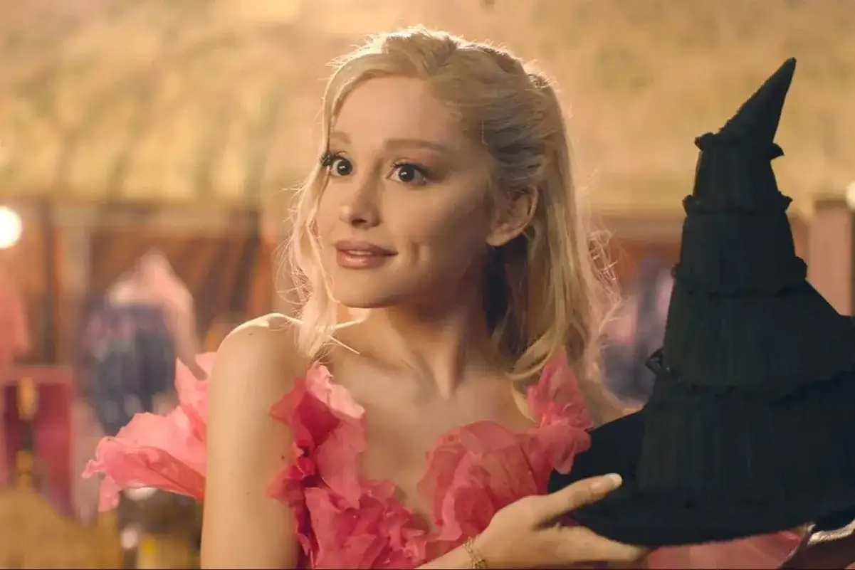 Ariana Grande w różowej sukni z kwiatami trzyma czarny kapelusz. Scena z jej filmów.