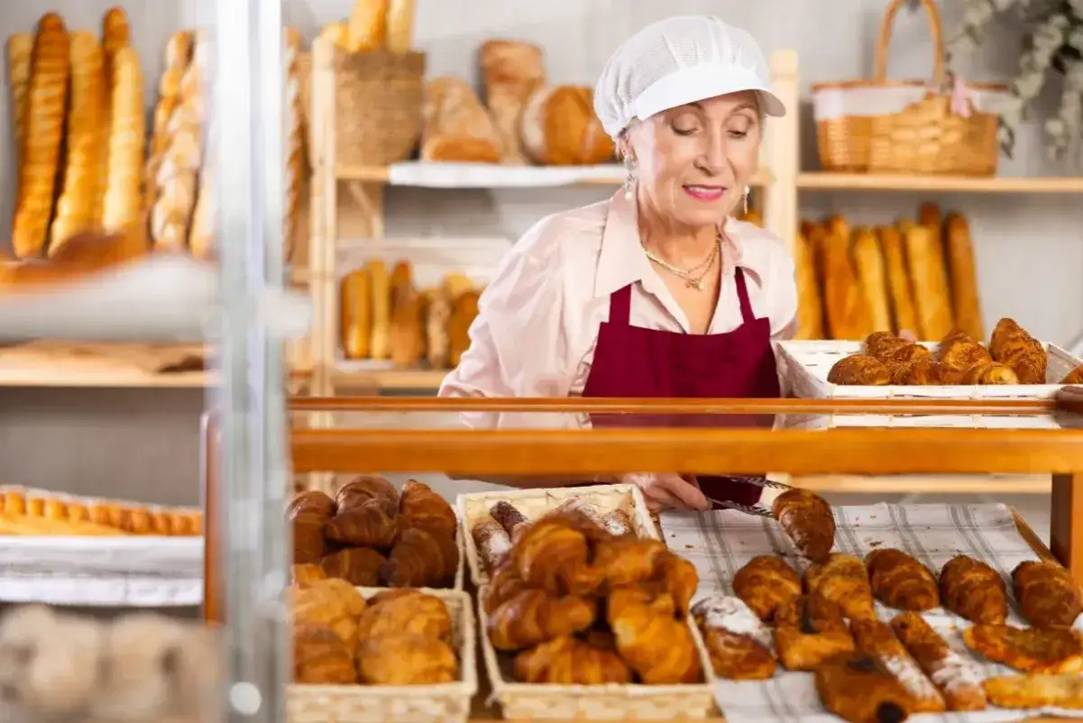 Starsza pani piekarz z uśmiechem układa świeże croissanty. To ulga dla pracujących seniorów, którzy wciąż mają pasję.