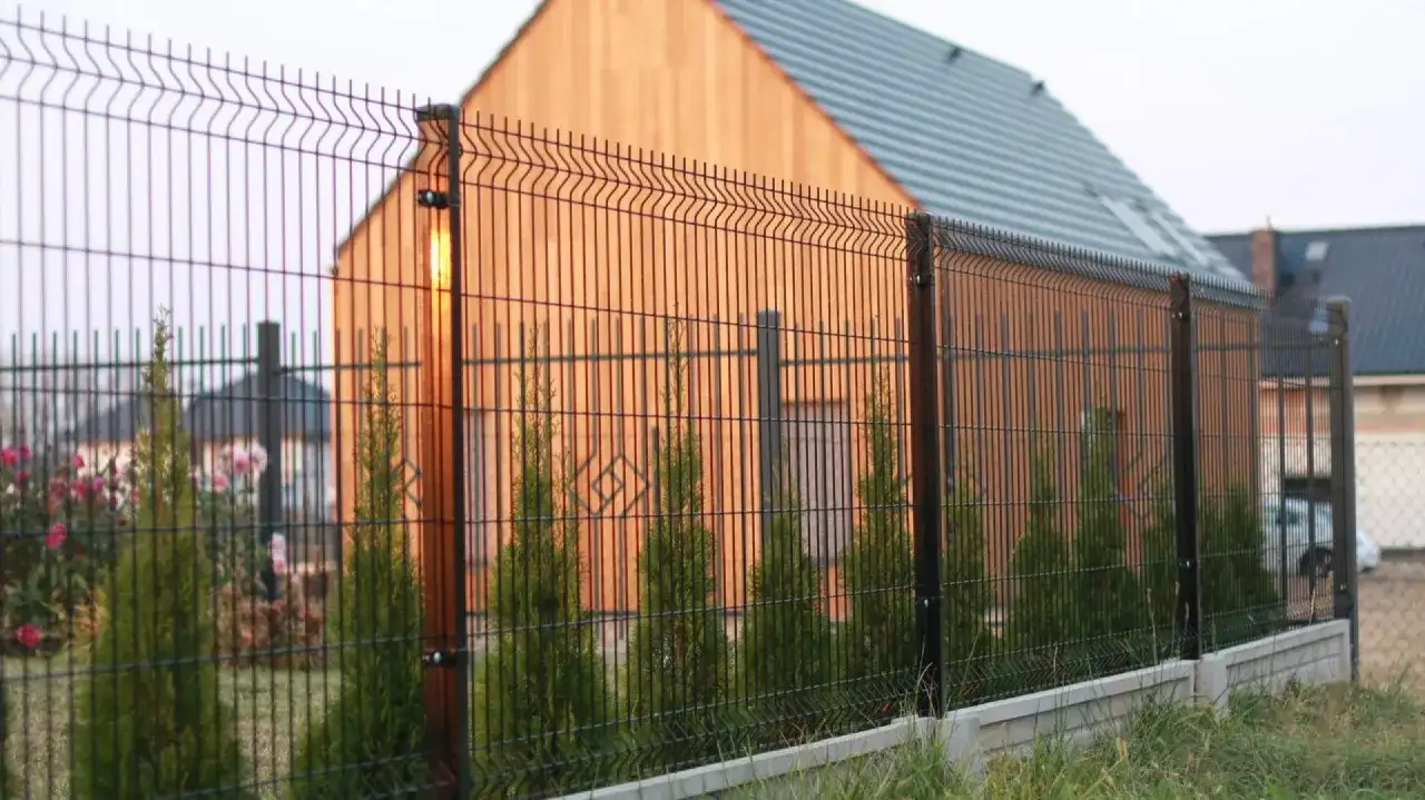 Nowy dom otoczony najprostszym ogrodzeniem. Wzdłuż siatki rosną tuje i kwitnące krzewy.