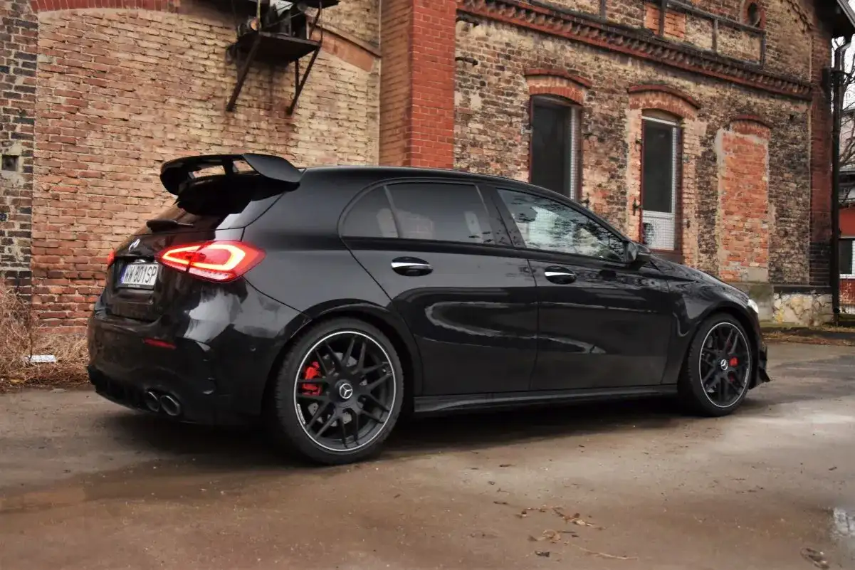 Czarny Mercedes-AMG A45s z masywnym spojlerem i czerwonymi zaciskami hamulcowymi stoi na mokrej nawierzchni przed starą ceglaną kamienicą.