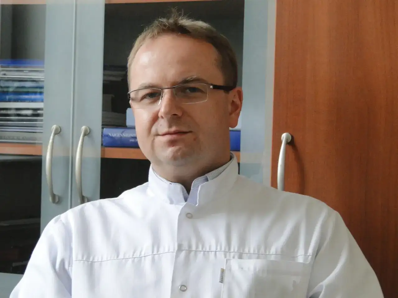 Portret mężczyzny w okularach i białym fartuchu lekarskim, na tle regałów z książkami.
