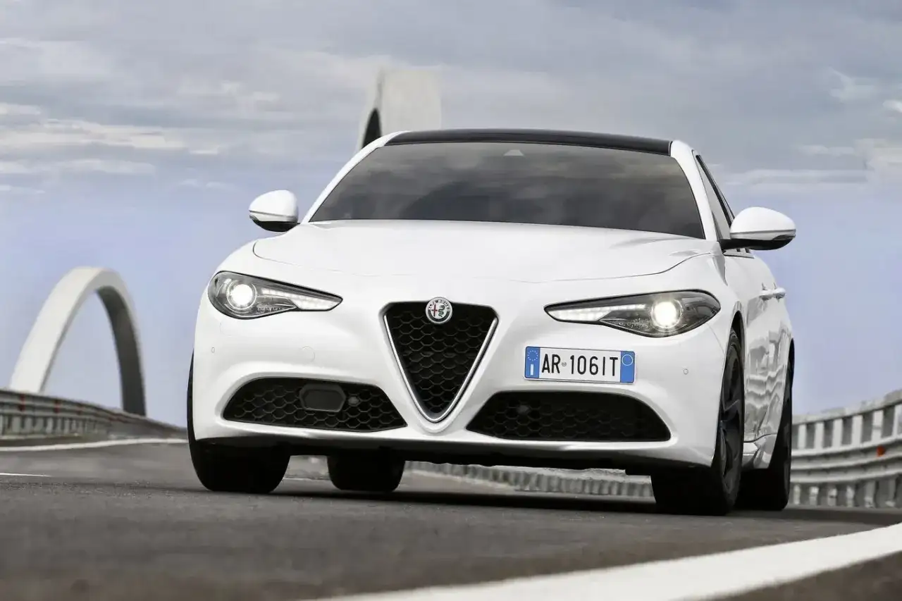 Biała Alfa Romeo Giulia Quadrifoglio z czarnym dachem pędzi po drodze z mostami w tle.