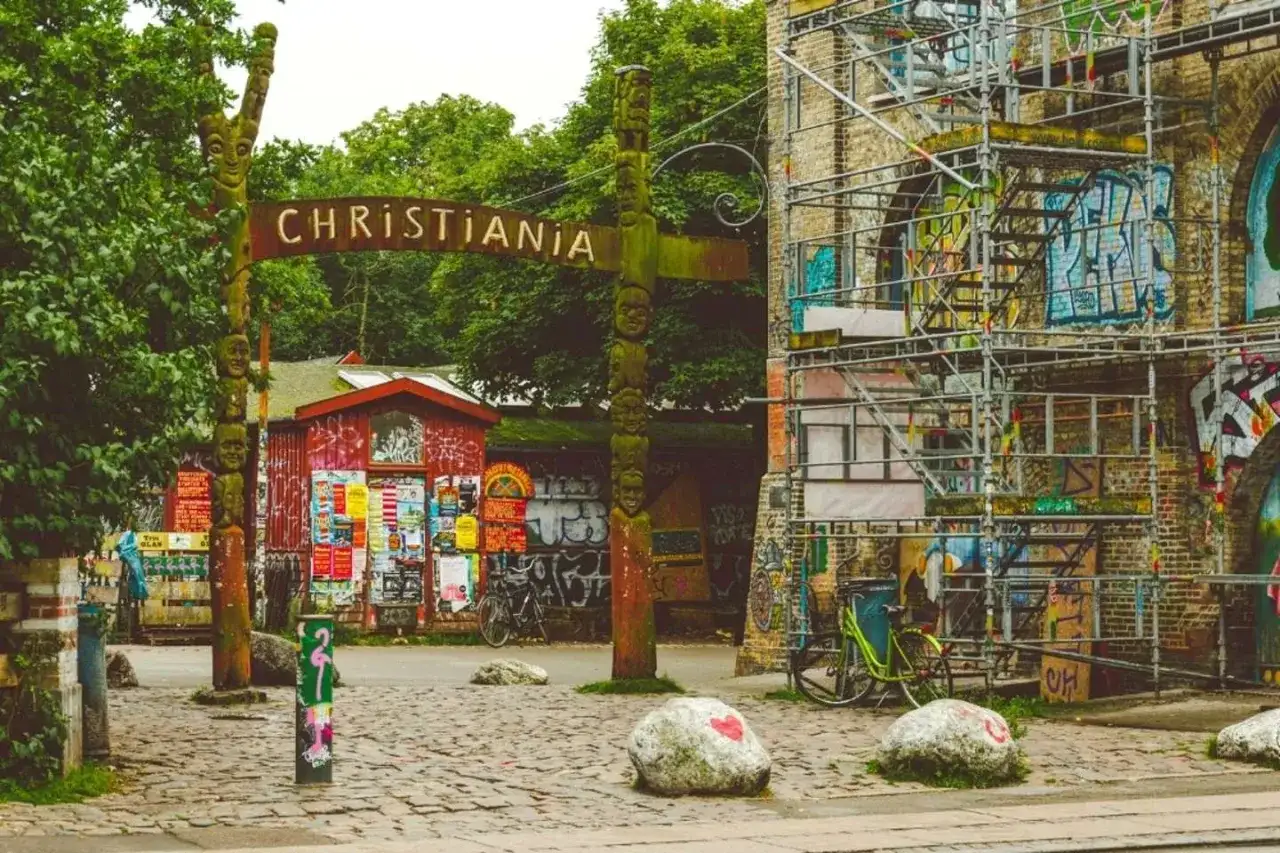 Drewniany totem i tablica z napisem "CHRISTIANIA" wita w tej wolnej dzielnicy. Obok czerwony budynek z plakatami i rowery.