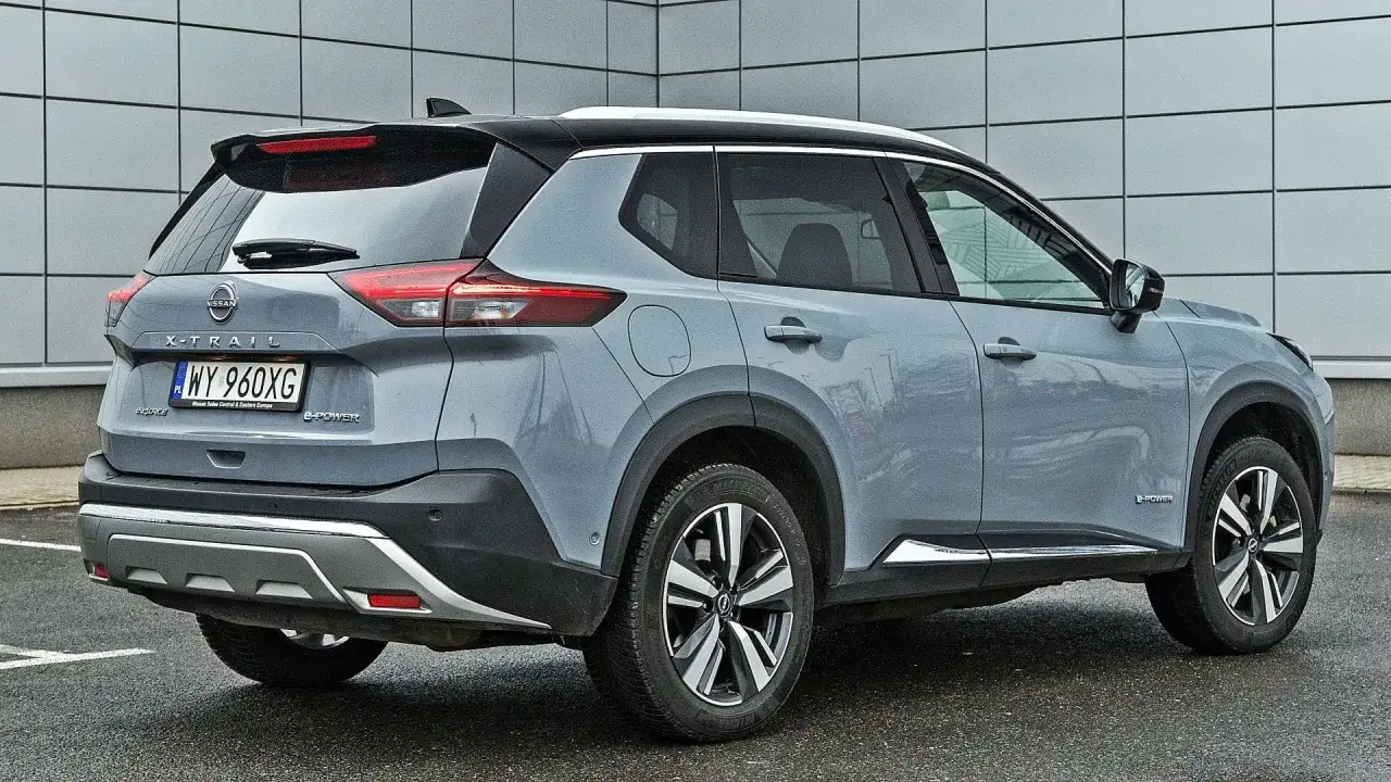 Szary Nissan X-Trail e-POWER z czarnym dachem i srebrnymi akcentami stoi na mokrym parkingu przed nowoczesnym budynkiem.