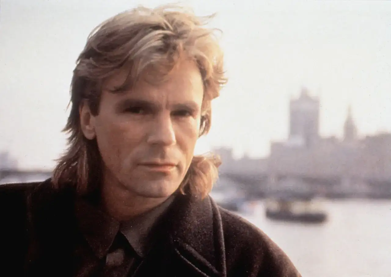 MacGyver obsada: Richard Dean Anderson w płaszczu, z charakterystyczną fryzurą, na tle zamglonego miasta.