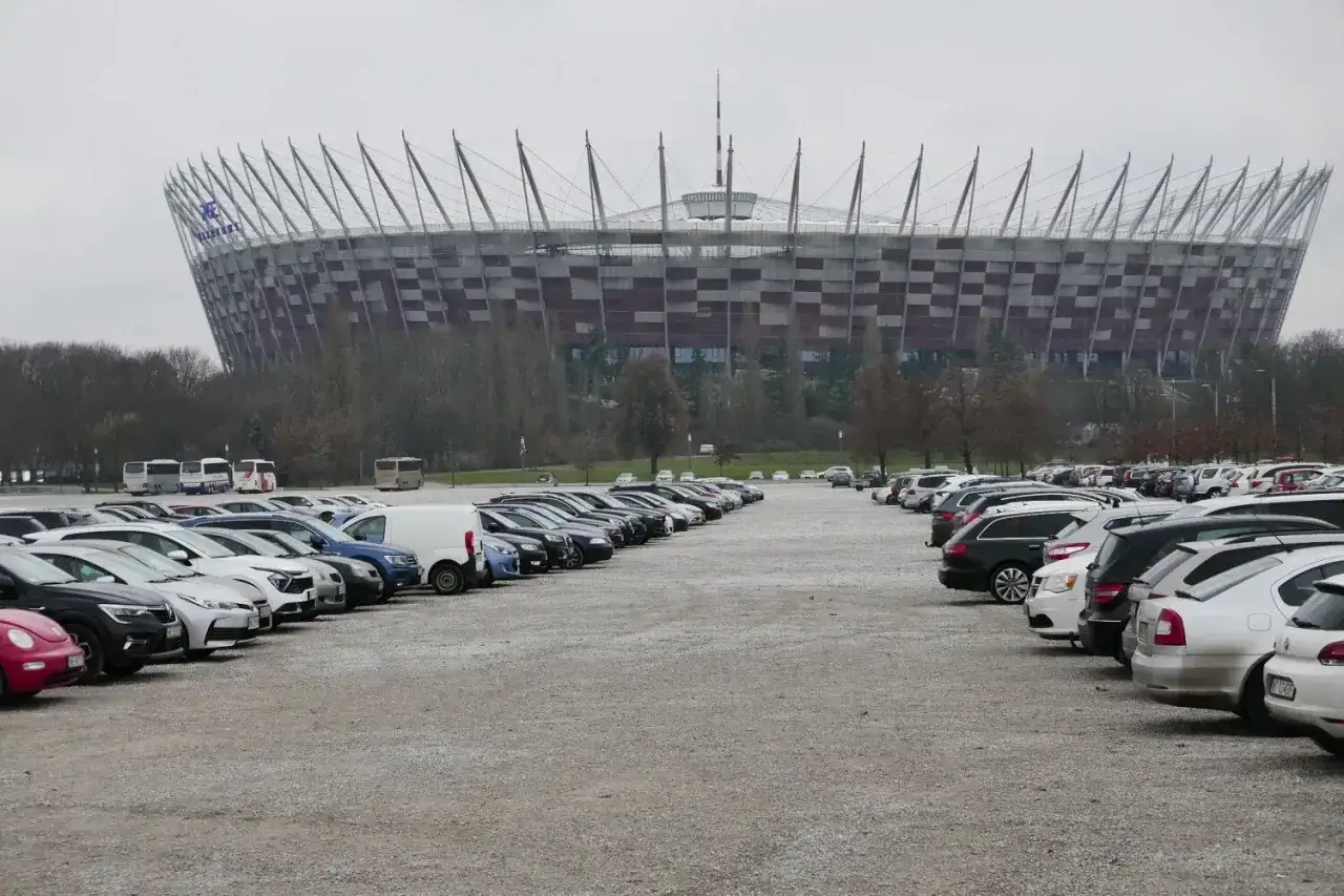 Duży parking przed Stadionem Narodowym w Warszawie. Widać rzędy samochodów i autobusów na błoniach stadionu.
