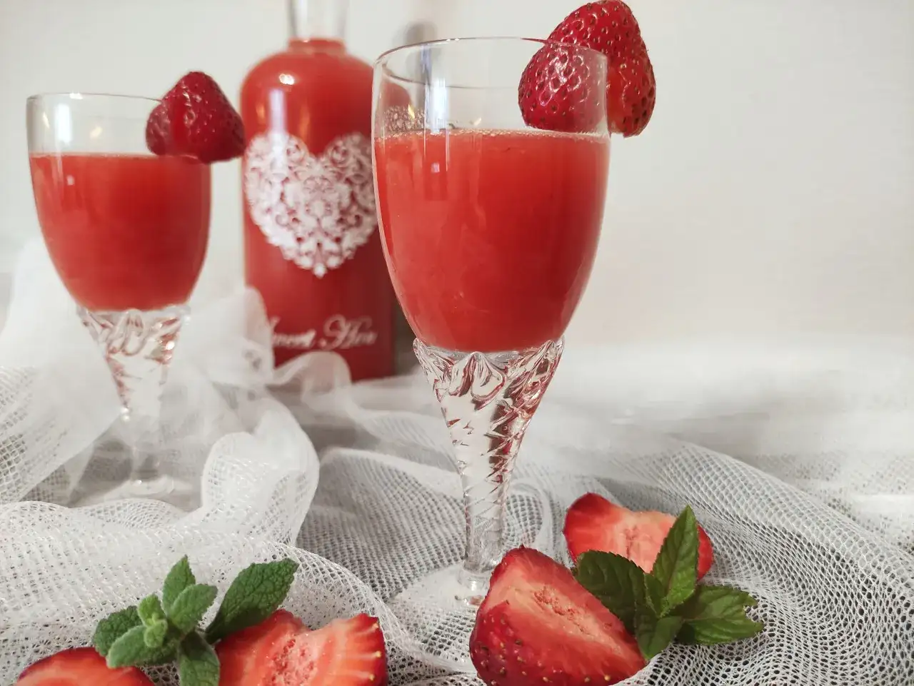Dwa kieliszki z orzeźwiającym, czerwonym drinkiem, ozdobione truskawkami. Obok butelka z napisem "Sweet Heart", sugerująca pyszny alkohol truskawkowy.