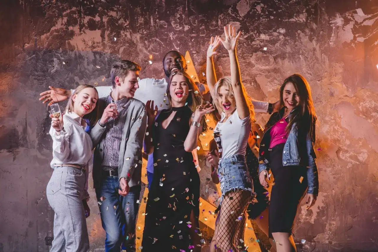 Grupa młodych ludzi świętuje, sypią się konfetti. Inspiracja, jak się ubrać do klubu: sukienka, jeansy, topy, marynarka.