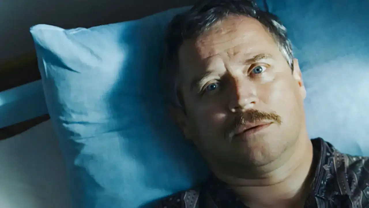 Mężczyzna z wąsami leży na niebieskiej poduszce, jego oczy patrzą w górę. Może to scena z filmu "Dzień świra", gdzie reżyser ukazuje codzienne zmagania bohatera.