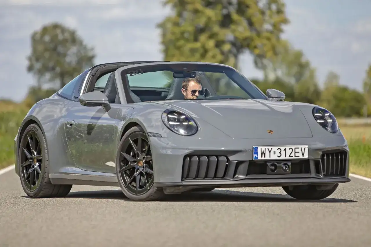 Szary Porsche 911 Carrera Targa z otwartym dachem, prowadzony przez mężczyznę w okularach przeciwsłonecznych, jedzie po asfaltowej drodze.