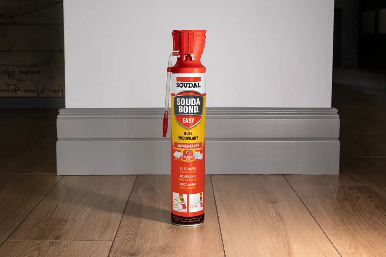 Puszka kleju budowlanego Soudal Easy Bond na drewnianej podłodze, obok szarej listwy przypodłogowej.