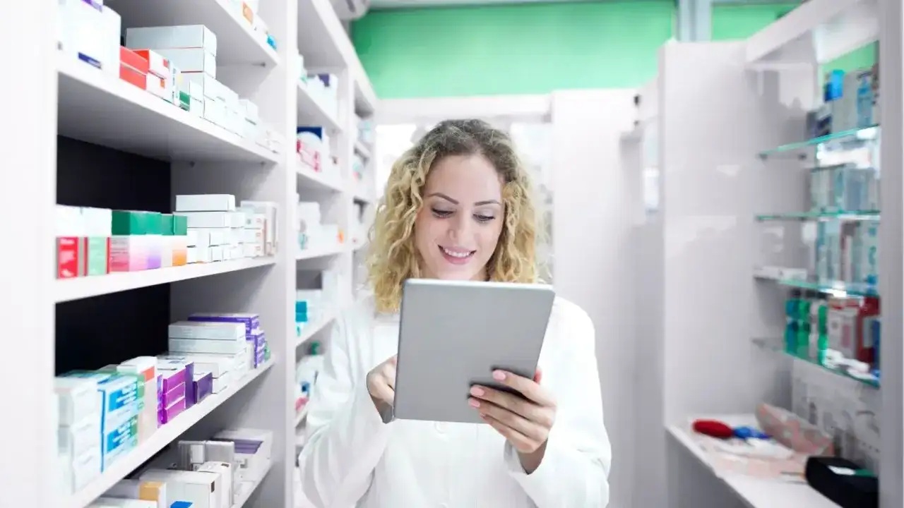 Farmacéutica sonriente revisa inventario en su tablet, rodeada de estantes llenos de productos de atida mifarma.
