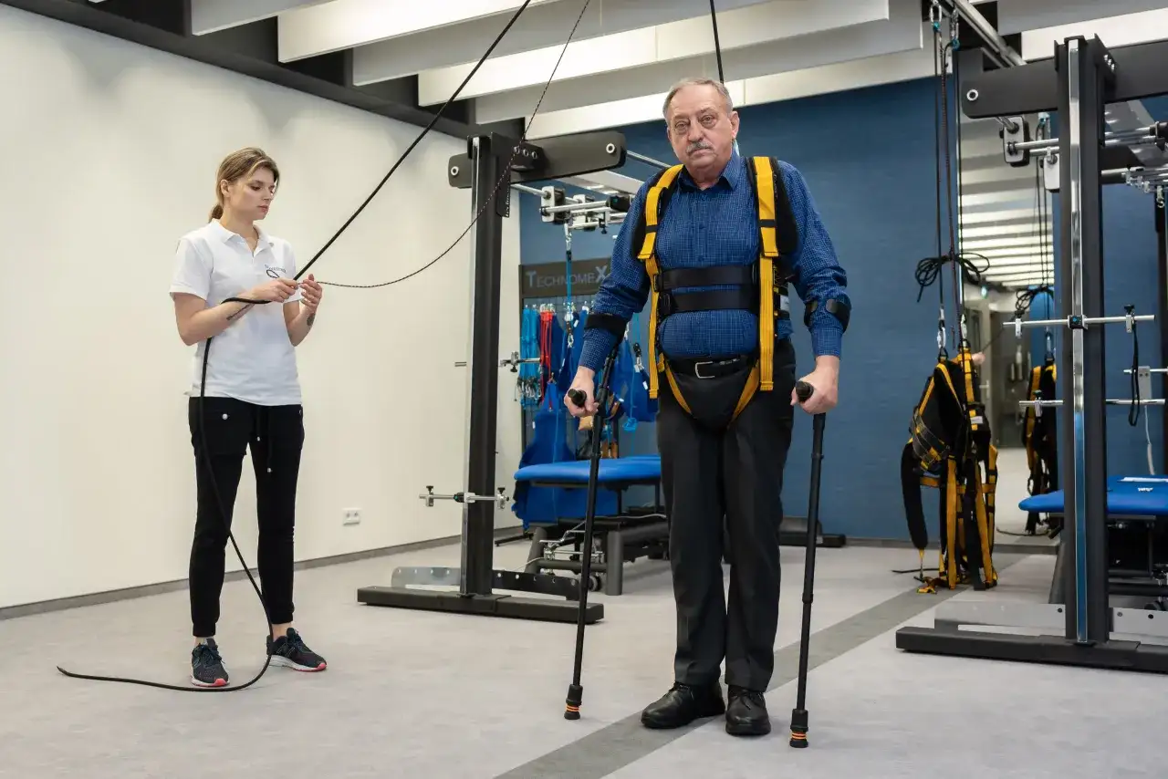 Starszy mężczyzna w specjalistycznej uprzęży i z kulami ćwiczy pod okiem fizjoterapeutki. To kluczowy etap rehabilitacji neurologicznej.