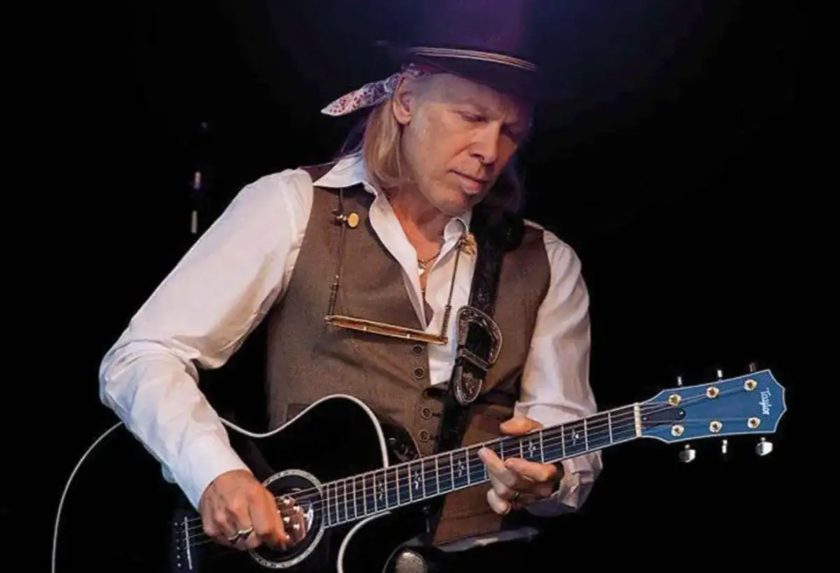 Elliott Murphy Zaragoza, con sombrero y chaleco, toca la guitarra acústica en un concierto.