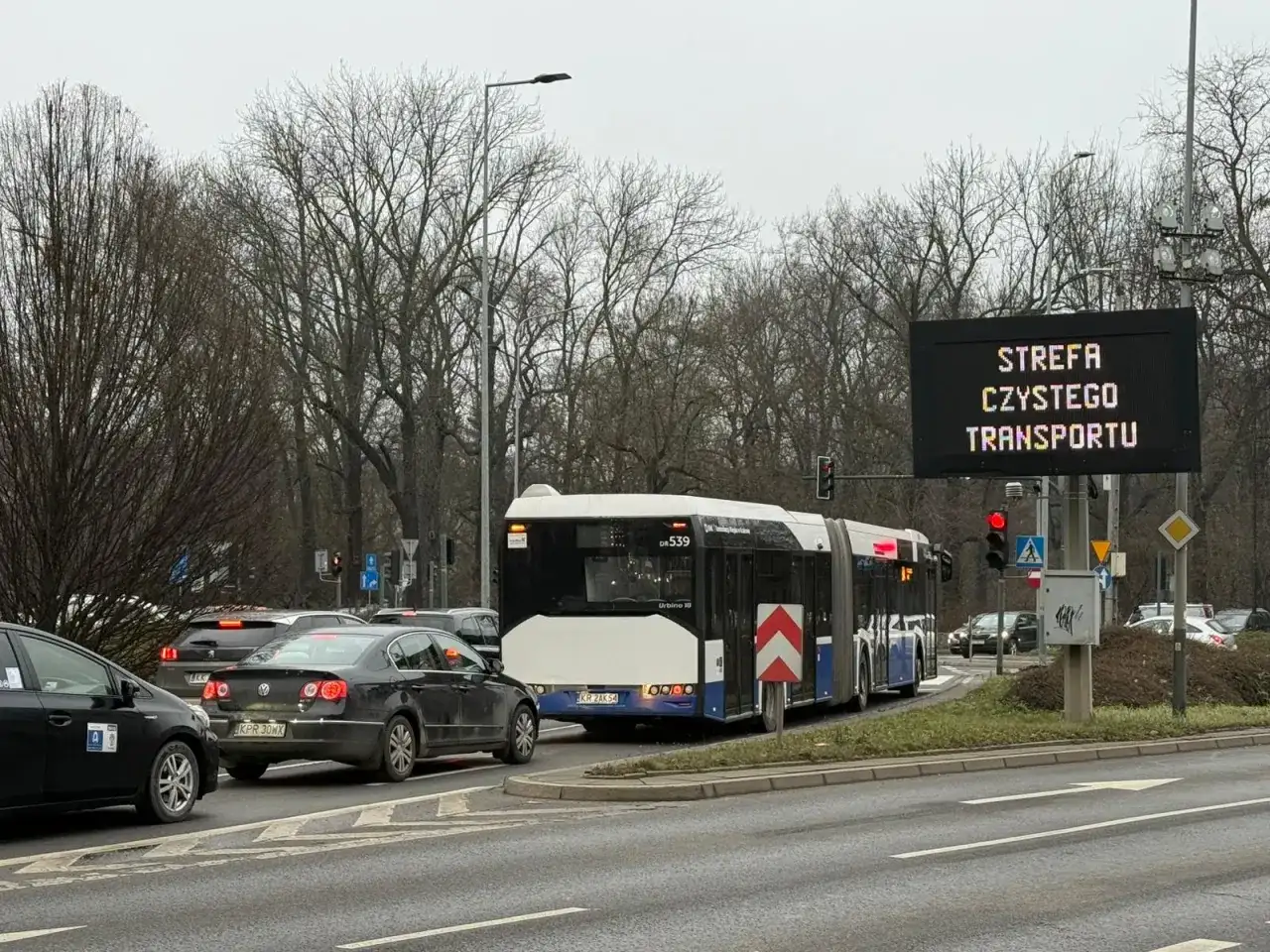 Autobus i samochody stoją w korku pod znakiem informującym o krakowskiej strefie czystego transportu.