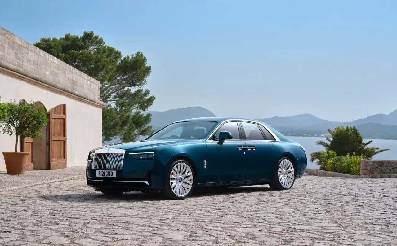 Elegancki Rolls-Royce Ghost w kolorze głębokiego turkusu zaparkowany na kamiennym dziedzińcu, z widokiem na morze i góry.