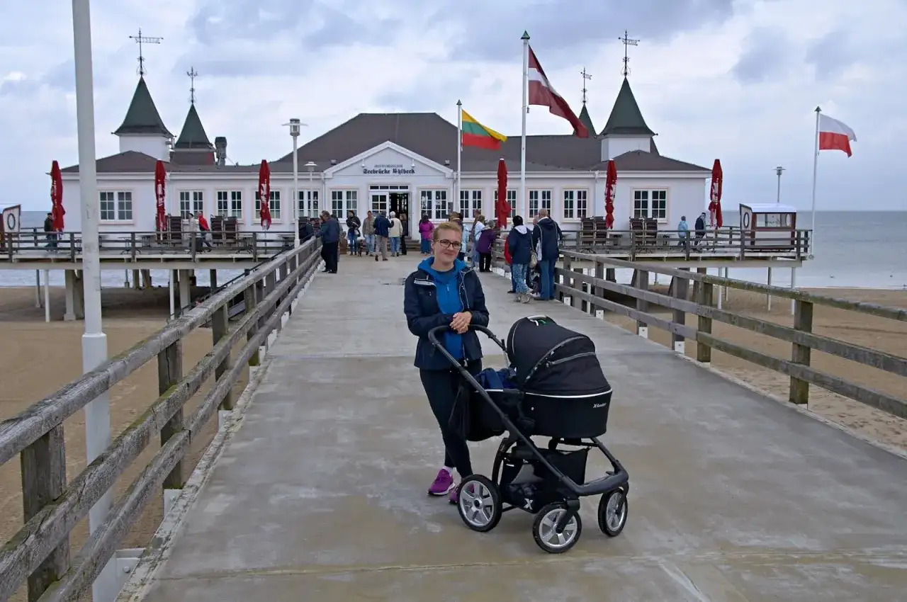 Kobieta z wózkiem na molo w Ahlbecku. W tle historyczny budynek i plaża, idealne miejsce na rodzinne atrakcje.